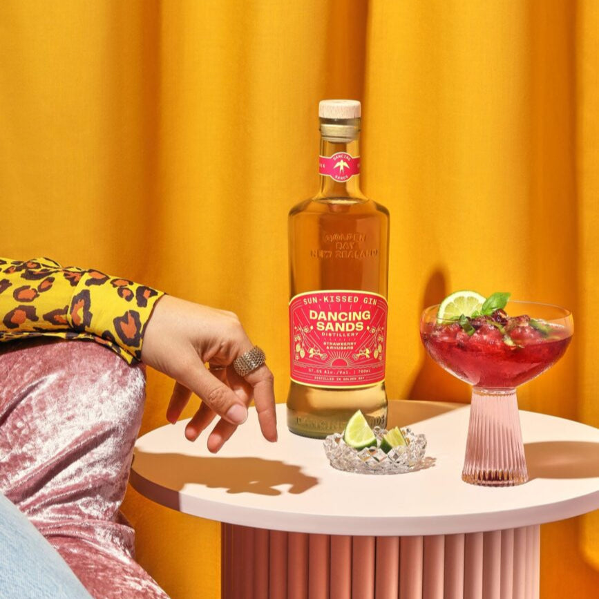 Load image into Gallery viewer, Bottle of 'Dancing Sands Sun Kissed Gin' on a small table with a cocktail and a person's hand in the background.