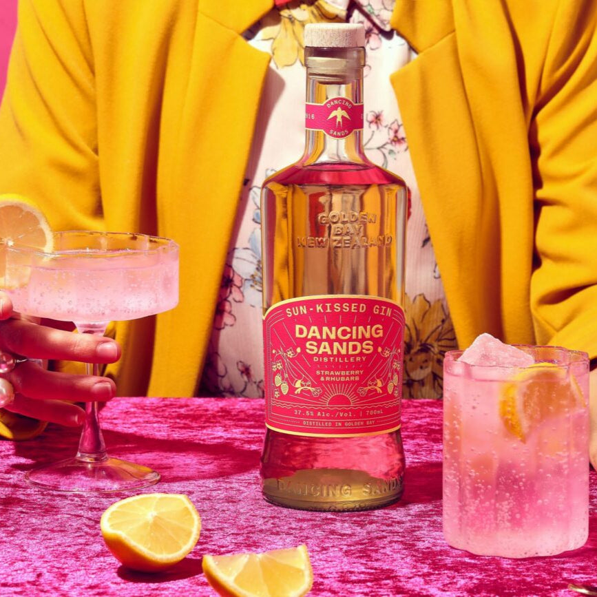 Person in a yellow jacket with a bottle of 'Dancing Sands' gin and pink cocktails on a pink background