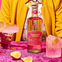 Load image into Gallery viewer, Person in a yellow jacket with a bottle of &#39;Dancing Sands&#39; gin and pink cocktails on a pink background