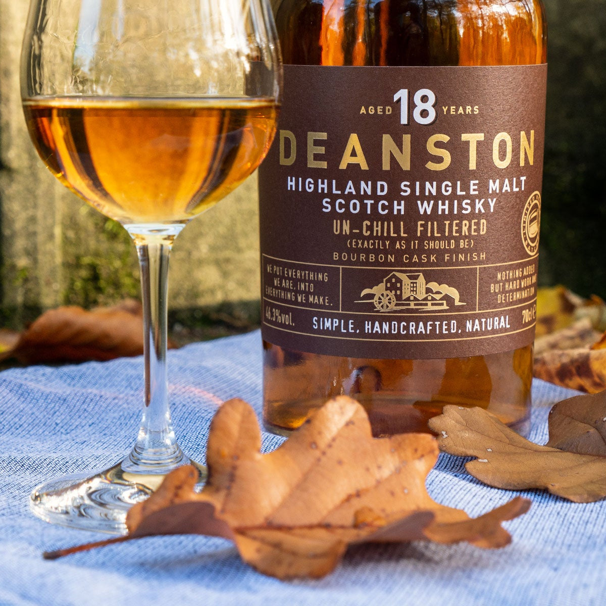 Load image into Gallery viewer, Deanston 18-year-old Scotch whisky bottle with a glass of whisky on a blue cloth with autumn leaves.