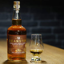 Load image into Gallery viewer, Deanston 18-year-old whisky bottle with a glass on a wooden surface against a dark brick wall.
