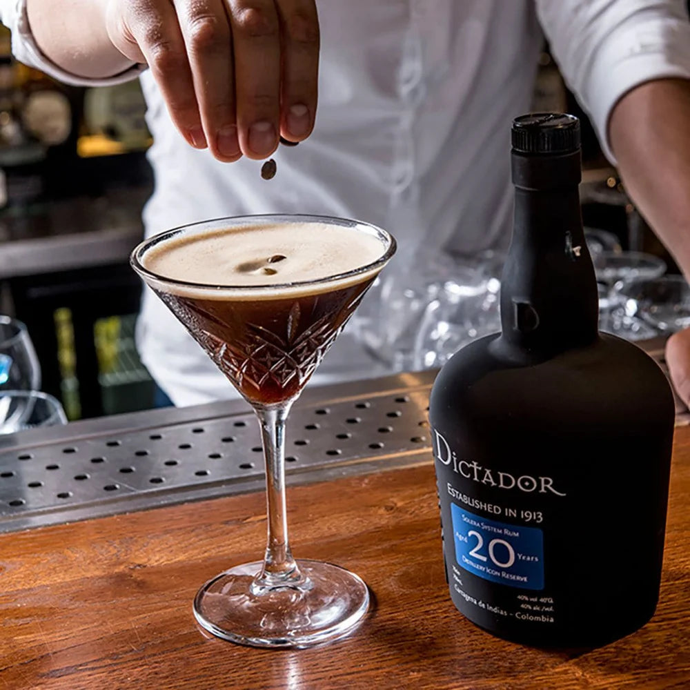 Person adding coffee beans to a cocktail glass with a bottle of Dictador 20 year old rum on a bar counter.