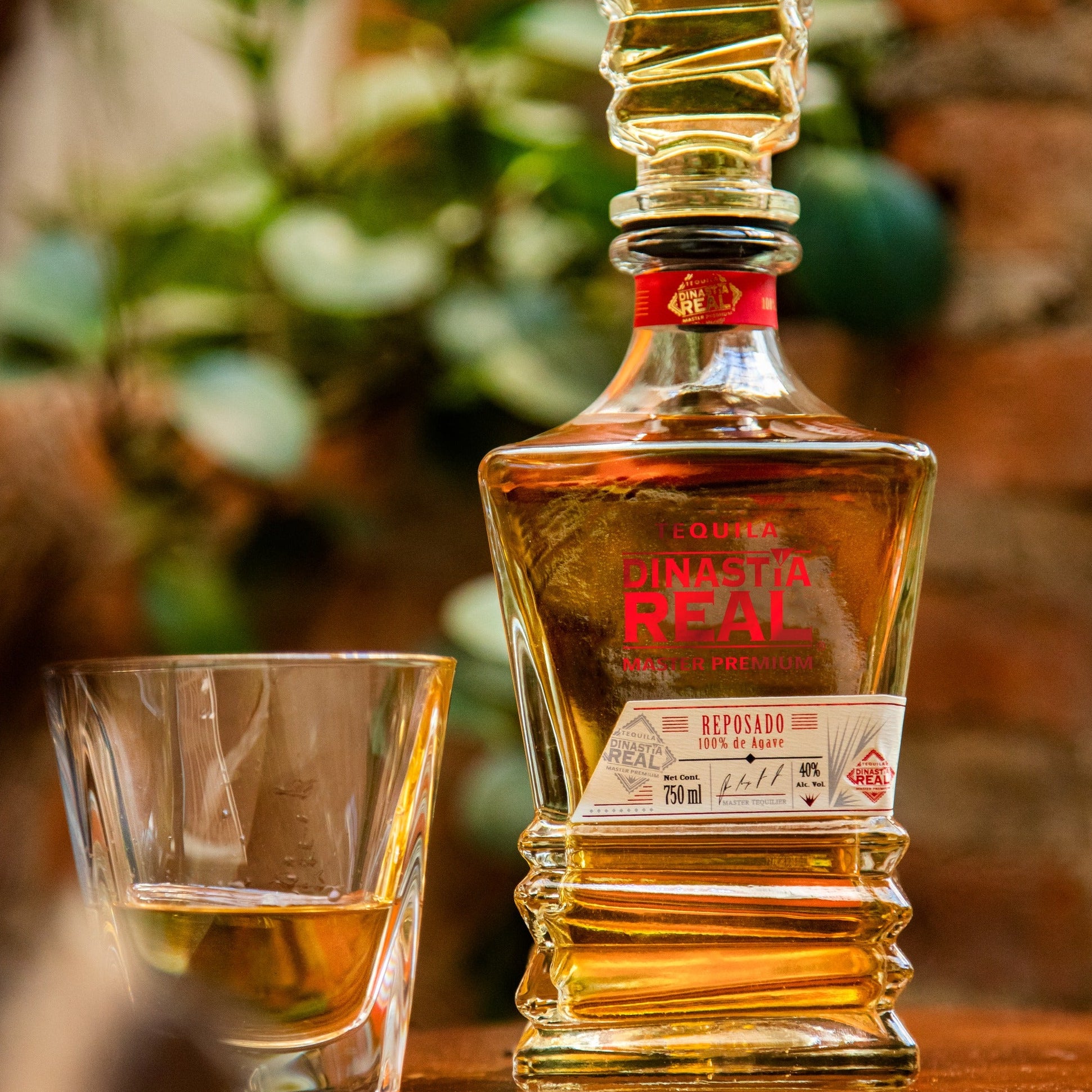 Load image into Gallery viewer, Bottle and glass of Dinastia Real Master Premium Reposado Tequila on a wooden surface with blurred greenery in the background.
