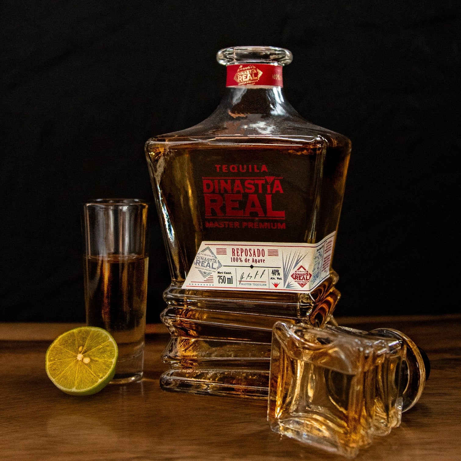 Load image into Gallery viewer, Dinastia Real Master Premium Reposado Tequila bottle with a glass and lime on a wooden surface, dark background.