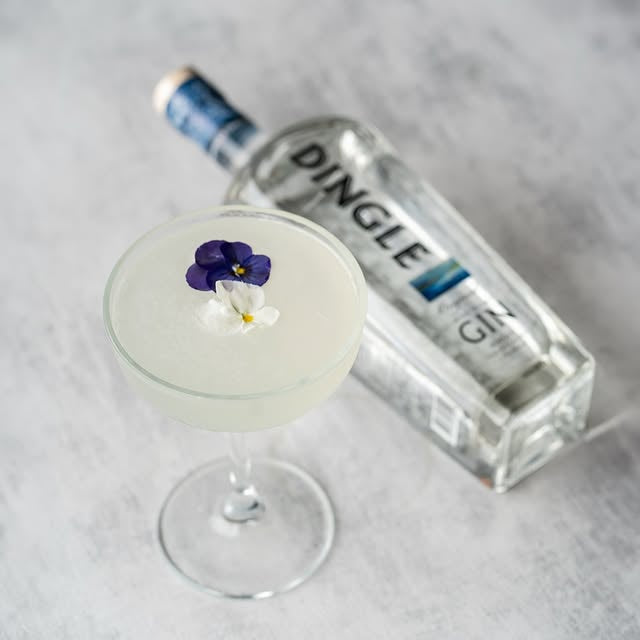 Cocktail with a floral garnish next to a bottle of Dingle gin on a gray surface