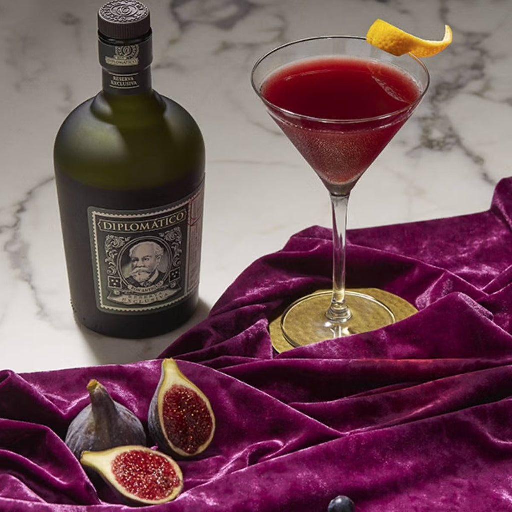 Bottle of Diplomatico Rum with a red cocktail and figs on a purple fabric