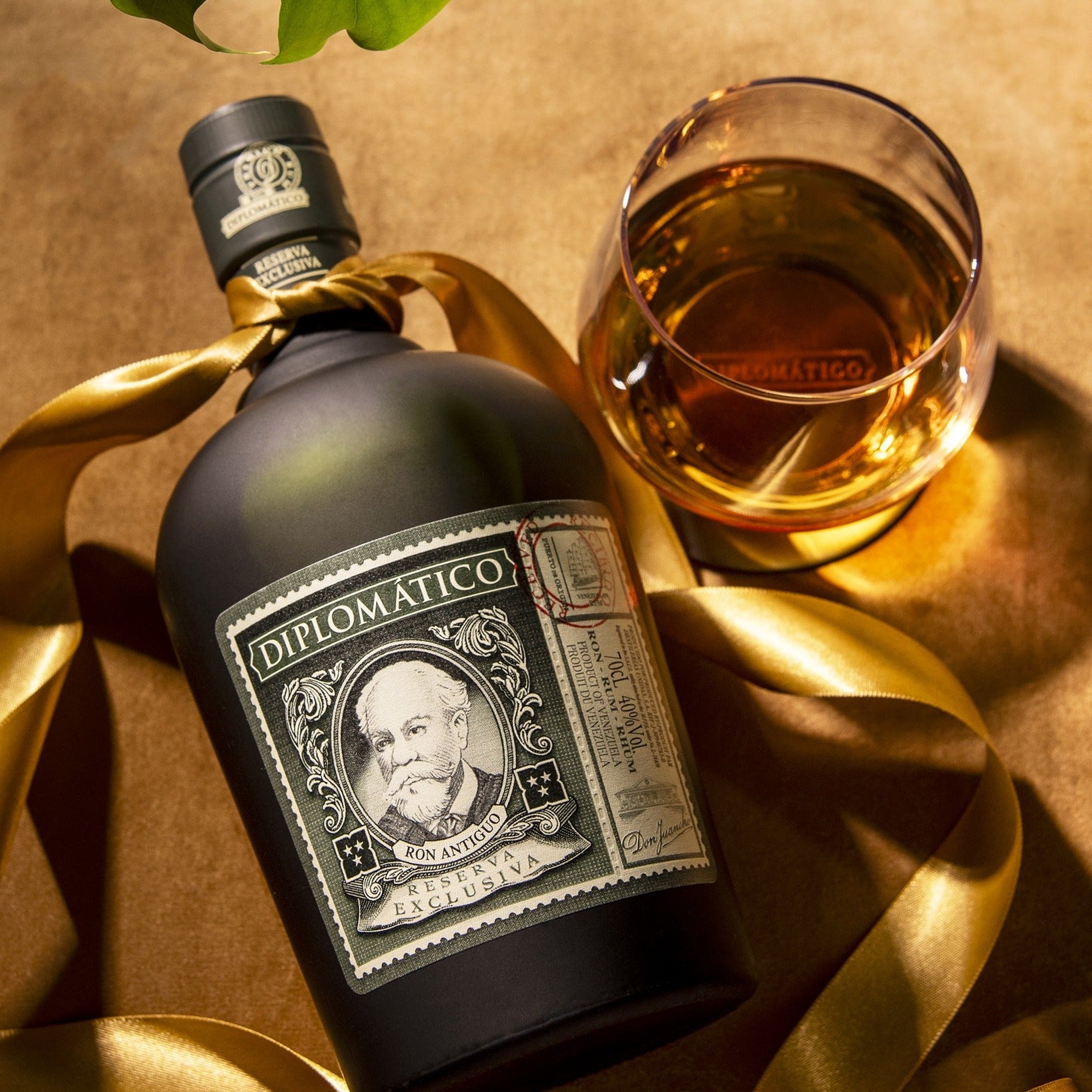 Load image into Gallery viewer, Diplomatico RESERVA EXCLUSIVA RUM bottle and glass on a gold ribbon with a brown background