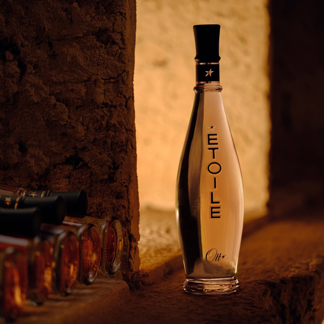 Load image into Gallery viewer, A clear glass bottle of Ott Etoile rosé wine with a black cap, standing on a rustic stone ledge in warm, dim lighting next to other bottles.