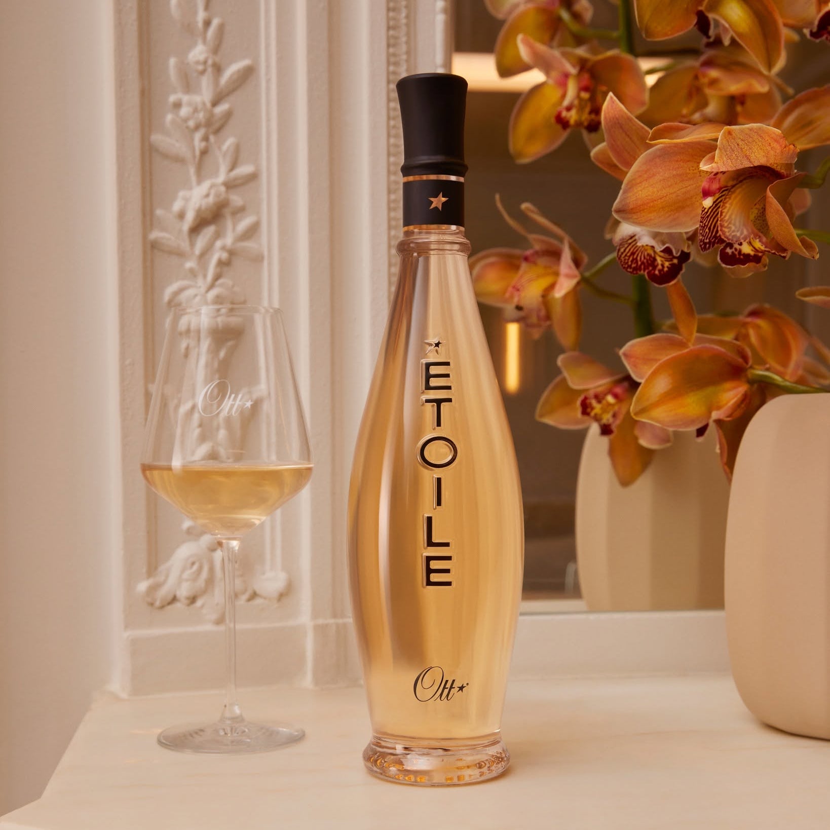 Load image into Gallery viewer, A bottle of Ott Etoile rosé wine with a glass of the pale golden wine and a vase of orange orchids in the background.