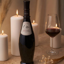 Load image into Gallery viewer, A bottle of red wine and some candles on a table.