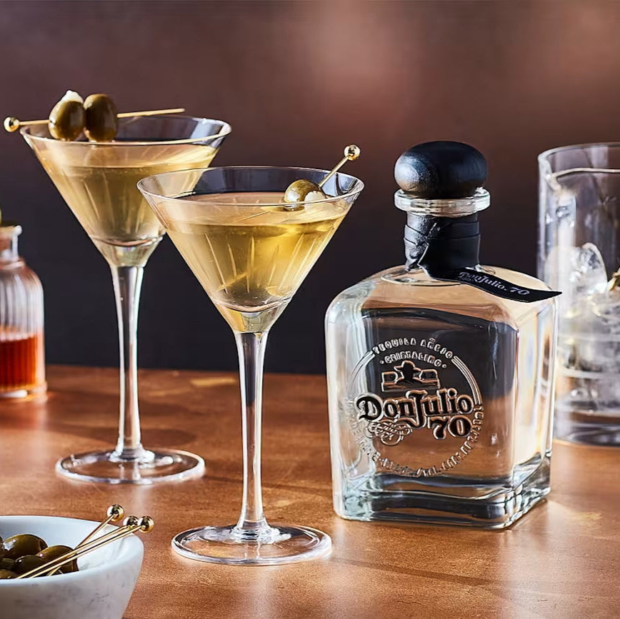 Two martini glasses with olive garnishes, a bottle of Don Julio 70 tequila, and a glass of water on a wooden surface.