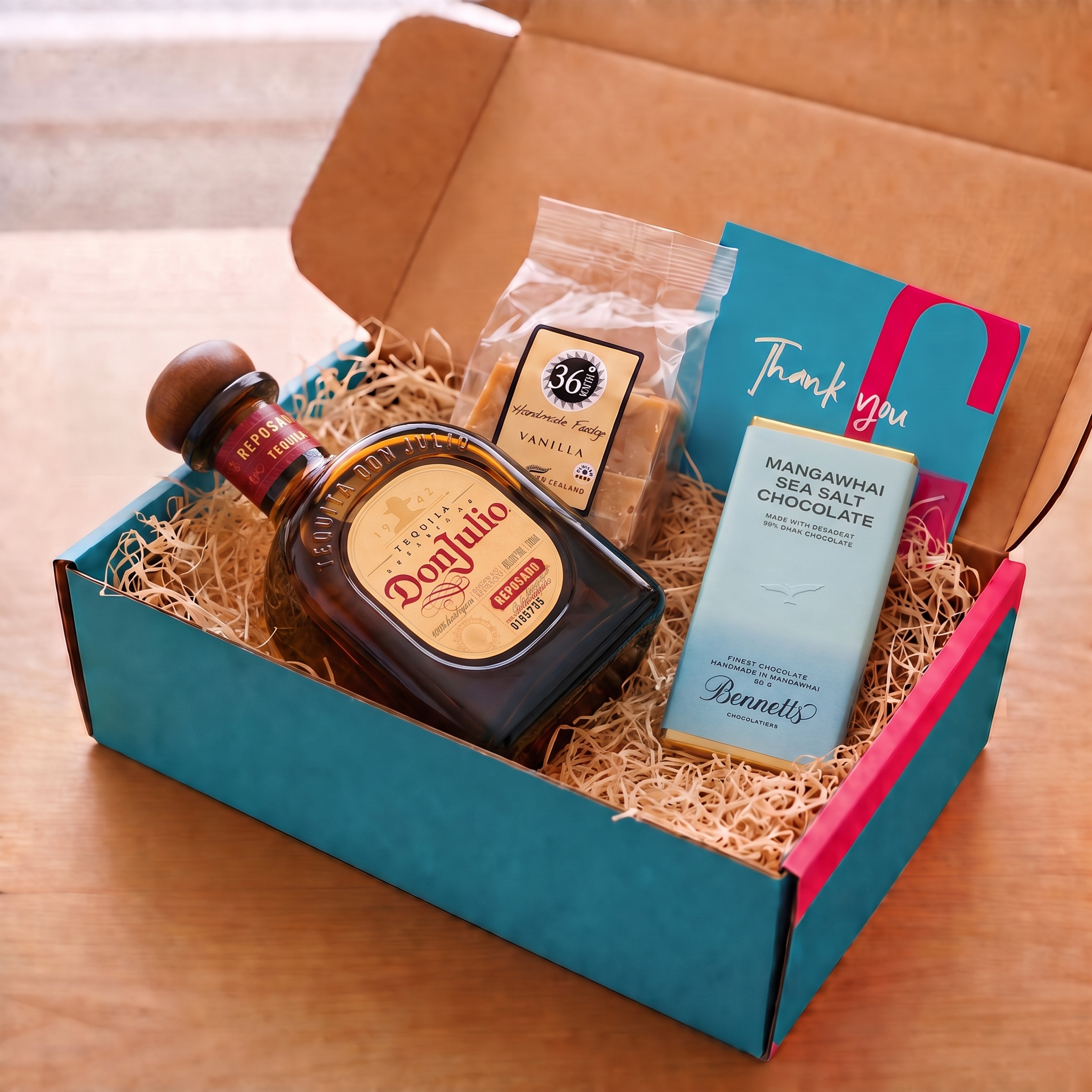 Load image into Gallery viewer, An open COVE 27 gift box filled with wood-wool and assorted treats, including a bottle of Don Julio reposado tequila, a bag of 36 Degrees Vanilla fudge, a Bennet's Mangawhai sea salt chocolate bar, and a blue and pink Thank You card, set on a wooden table.