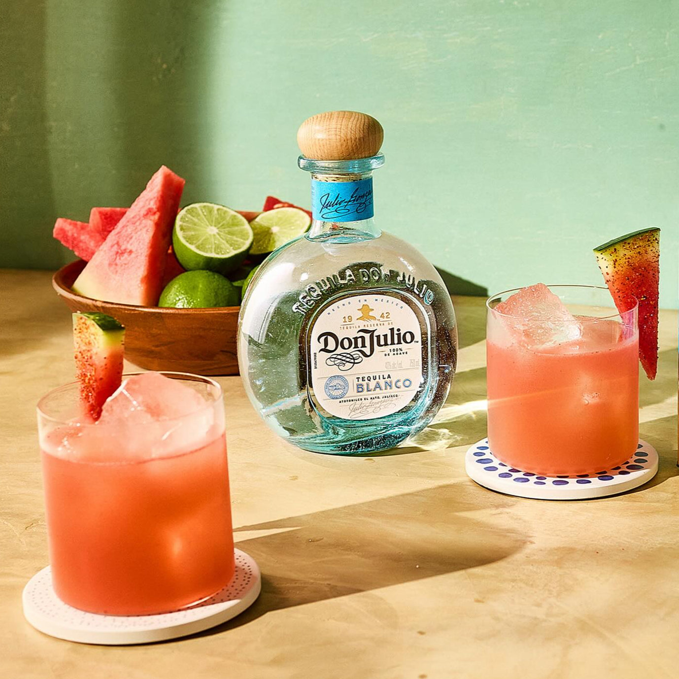Load image into Gallery viewer, Two glasses of pink cocktails with watermelon slices, a bottle of Don Julio tequila, and a bowl of fruit on a wooden surface.