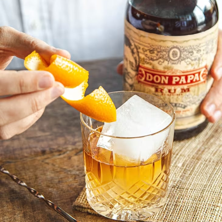 Glass of rum with ice and orange peel, next to a bottle of Don Papa Rum on a wooden surface.