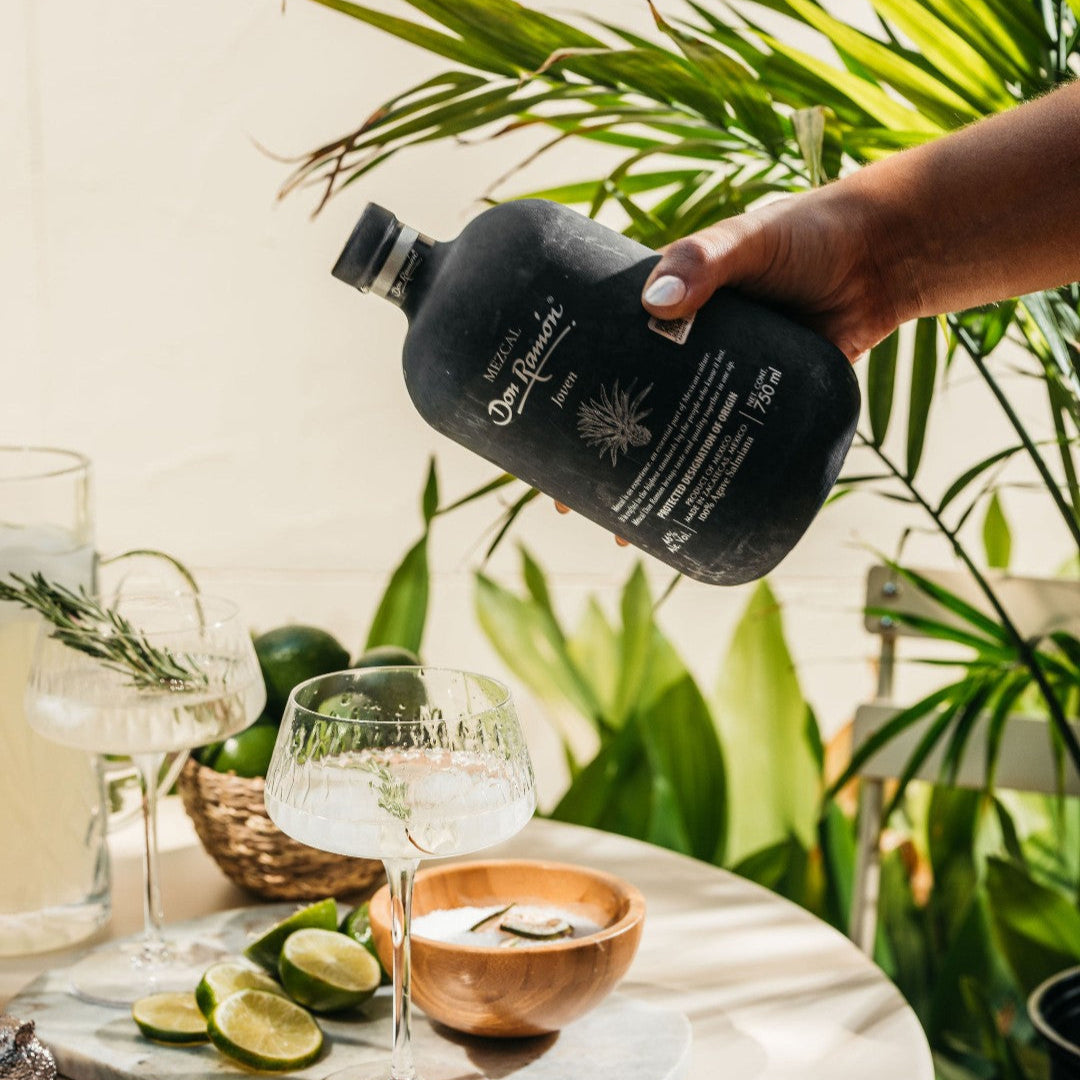 Load image into Gallery viewer, Person pouring a drink from a Don Ramon Mezcal black bottle into a glass on a table with plants in the background