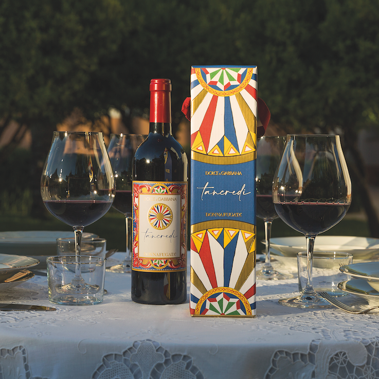 Load image into Gallery viewer, Donnafugata Tancredi Dolce Gabbana red wine bottle with colourful label and box, surrounded by glasses of red wine on a table outdoors.