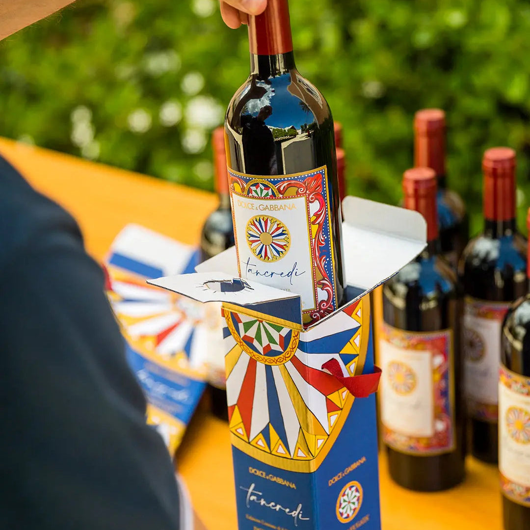 Load image into Gallery viewer, Person holding a bottle of Donnafugata Tancredi Dolce Gabbana red wine with decorative label, surrounded by more bottles on a table outdoors.
