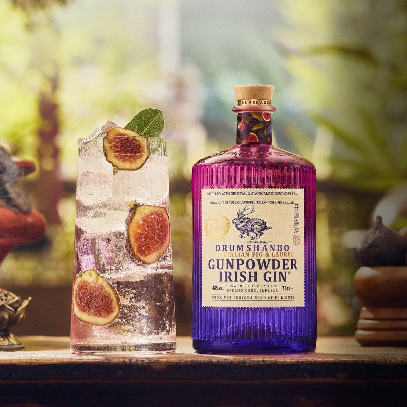 Load image into Gallery viewer, A bottle of Drumshambro Gunpowder Laurel &amp; Fig Irish Gin stands on a wooden surface beside a glass of gin with ice, fig slices, and mint, with blurred botanical props in the background.