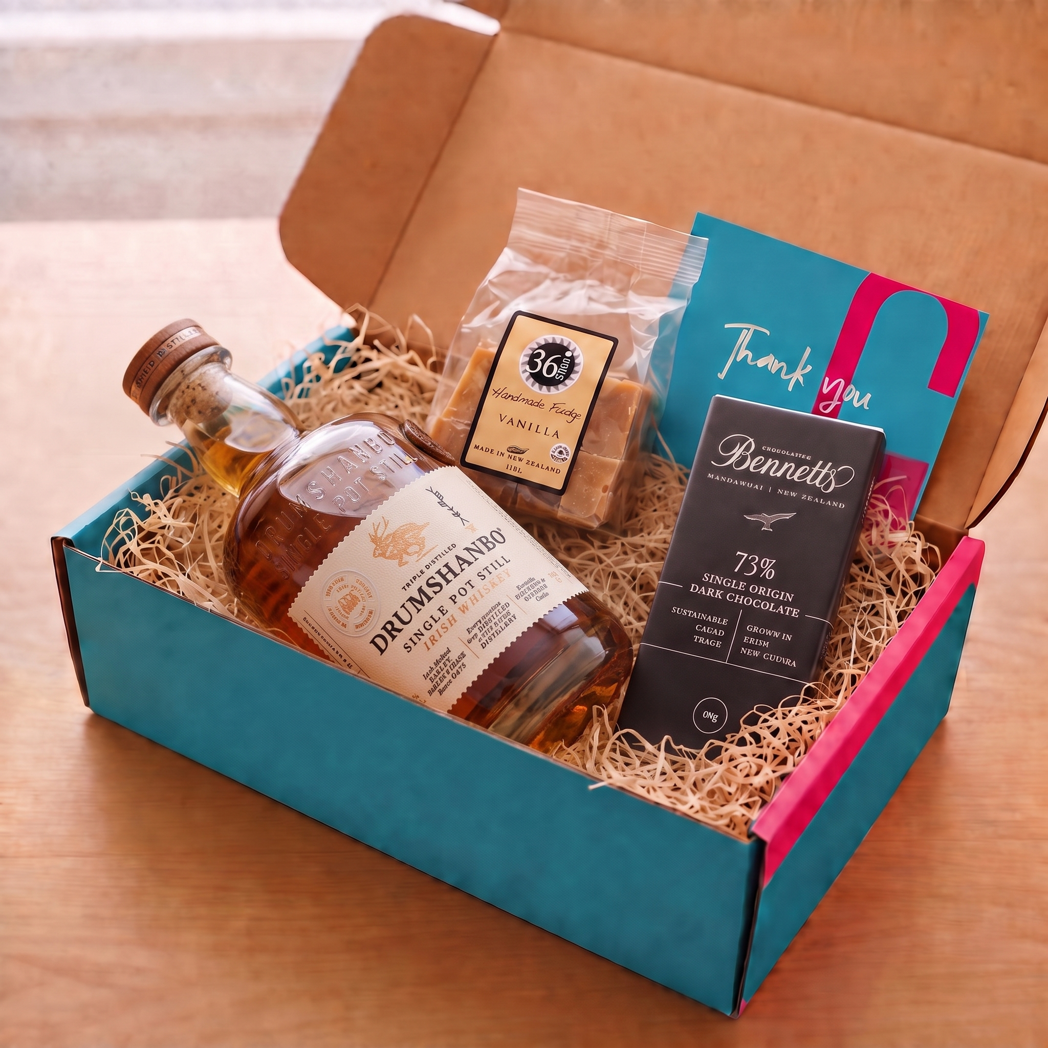 Load image into Gallery viewer, A gift box set on a wooden table containing a bottle of Drumshanbo Single Pot Still Irish Whiskey, a bag of 36 Degrees Vanilla fudge, a Bennett 73% Single Origin Dark Chocolate bar, and a teal and pink Thank You card inside a COVE 27 gift box with a turquoise and pink inner box filled with wood-wool.