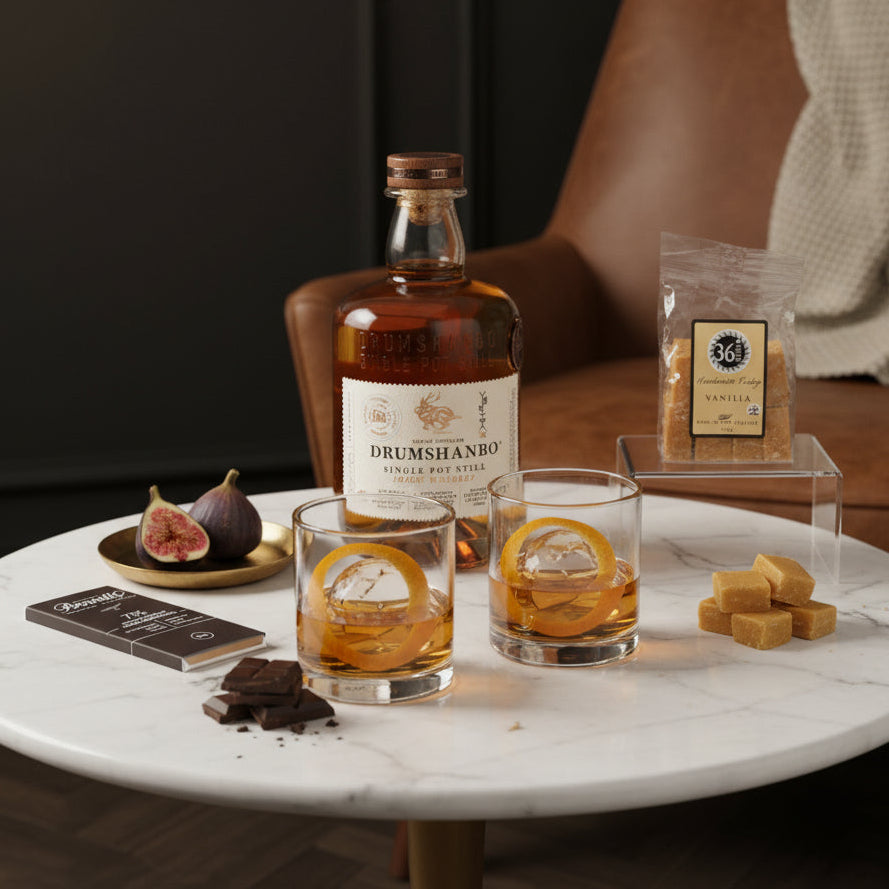 Load image into Gallery viewer, A styled tabletop scene with a bottle of Drumshanbo Single Pot Still Irish Whiskey, two clear glasses with orange slices and ice, a bag of 36 Degrees vanilla fudge, chocolate pieces, figs, and a Bennetts dark chocolate bar on a white marble round table. A brown leather armchair with a cream throw is in the background against a dark panelled wall.