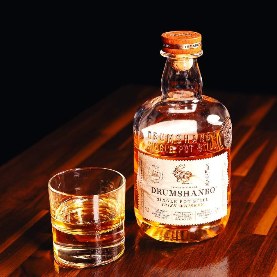 Load image into Gallery viewer, A bottle of Drumshanbo Single Pot Still Irish Whiskey with a small glass of the amber liquid beside it, set on a dark wooden table against a black background.
