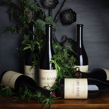Load image into Gallery viewer, Bottles of Dry-River-Pinot-Noir wine with green leaves on a wooden surface against a dark background