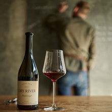 Load image into Gallery viewer, Dry-River-Pinot-Noir wine bottle and glass on a wooden table with a blurred person in the background