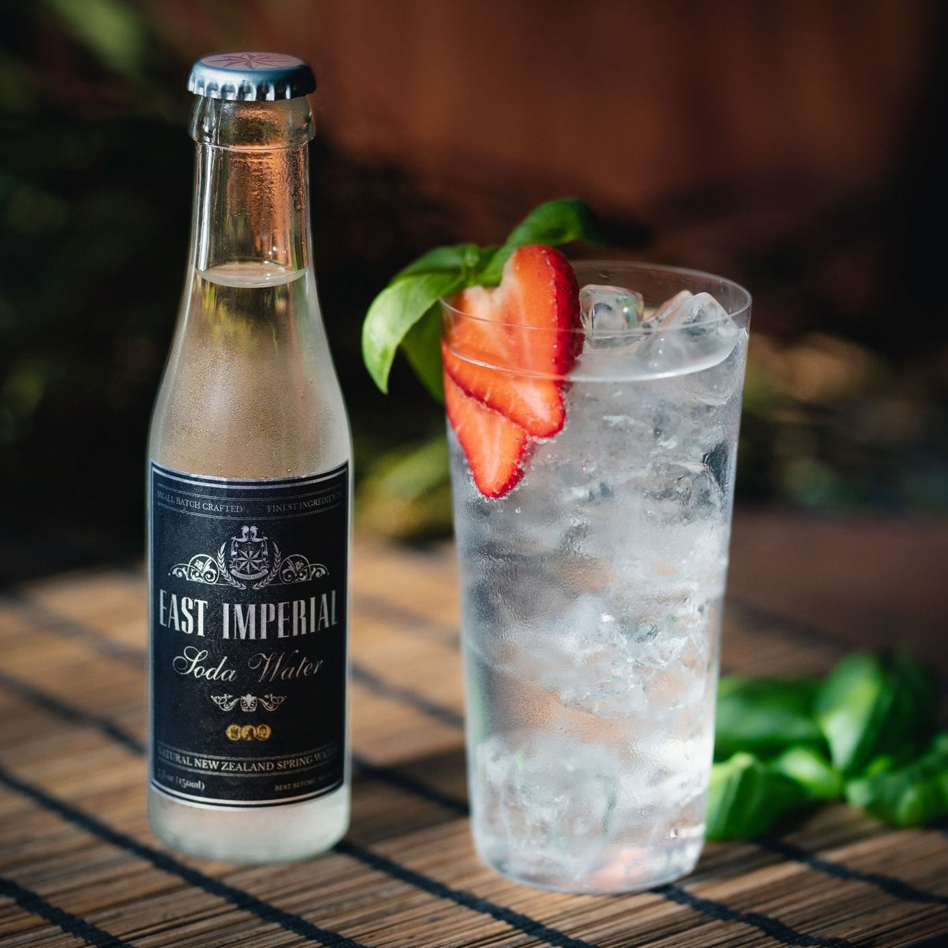 Load image into Gallery viewer, Bottle of East Imperial Soda Water next to a glass with ice and strawberries on a woven mat.