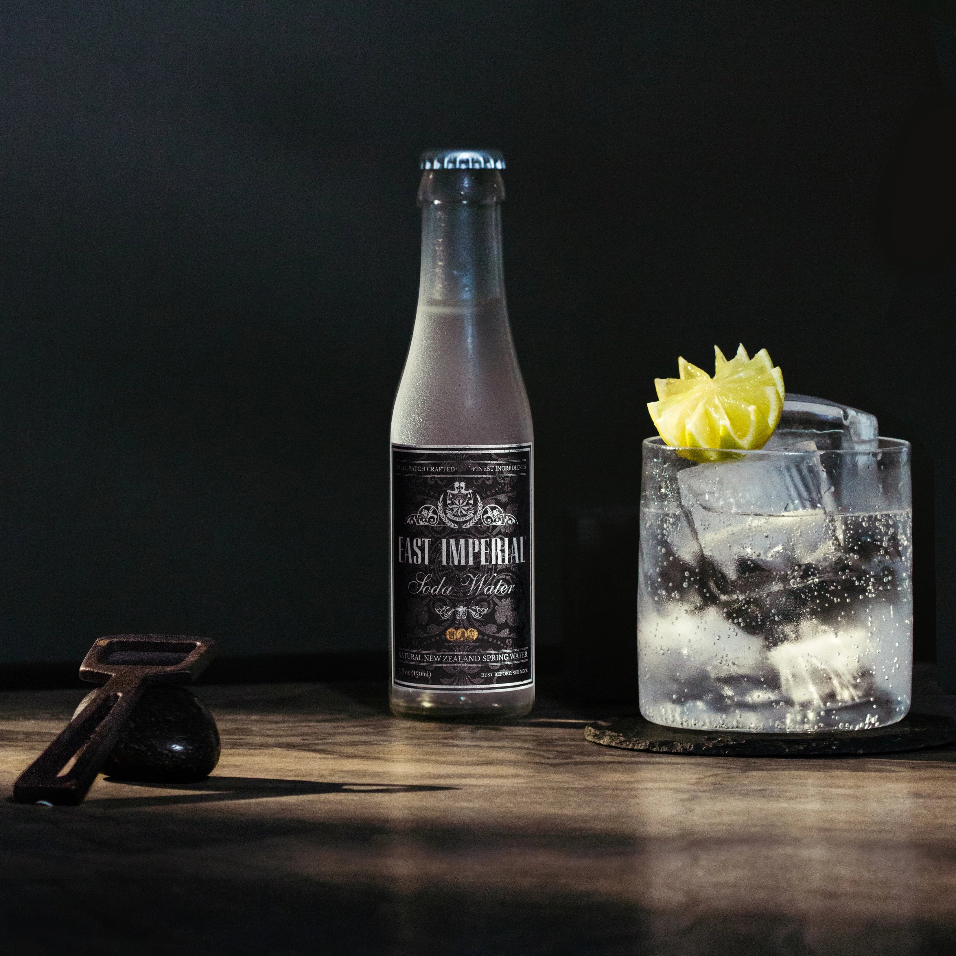 Load image into Gallery viewer, Bottle of East Imperial Soda Water and a cocktail with ice and a lemon twist on a dark background