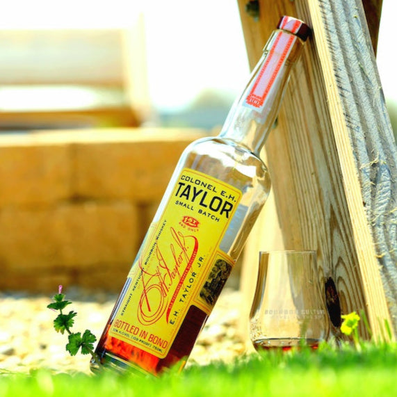 A bottle of EH-Taylor-Small-Batch-Kentucky-Straight-Bourbon-Whiskey next to tasting glass, outside on grass