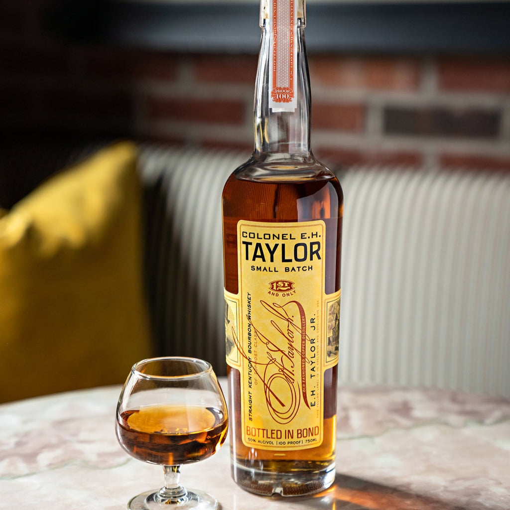 Bottle of Colonel E.H. Taylor whiskey with a glass on a marble surface, with a blurred background.