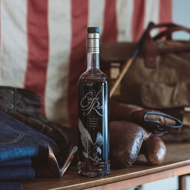 Load image into Gallery viewer, Bottle of whiskey labeled 'Eagle Rare' on a wooden surface with a flag and leather goods in the background.