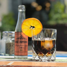 Load image into Gallery viewer, Bottle of East Imperial Grapefruit Tonic with a glass of soda and orange slice on a table outdoors.