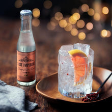 Load image into Gallery viewer, Bottle of East Imperial Grapefruit Tonic Water next to a cocktail with ice and lemon on a wooden tray.