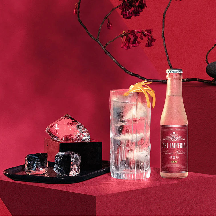 Load image into Gallery viewer, A bottle of East Imperial Tonic Water with a glass of tonic water and ice cubes on a red background.