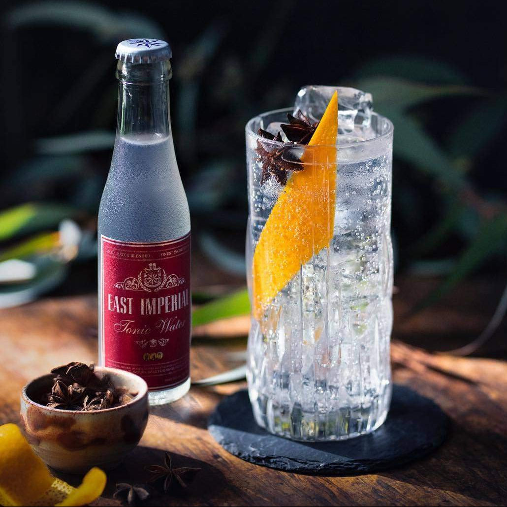 Load image into Gallery viewer, Bottle of East Imperial Tonic Water next to a cocktail with an orange slice on a wooden surface.