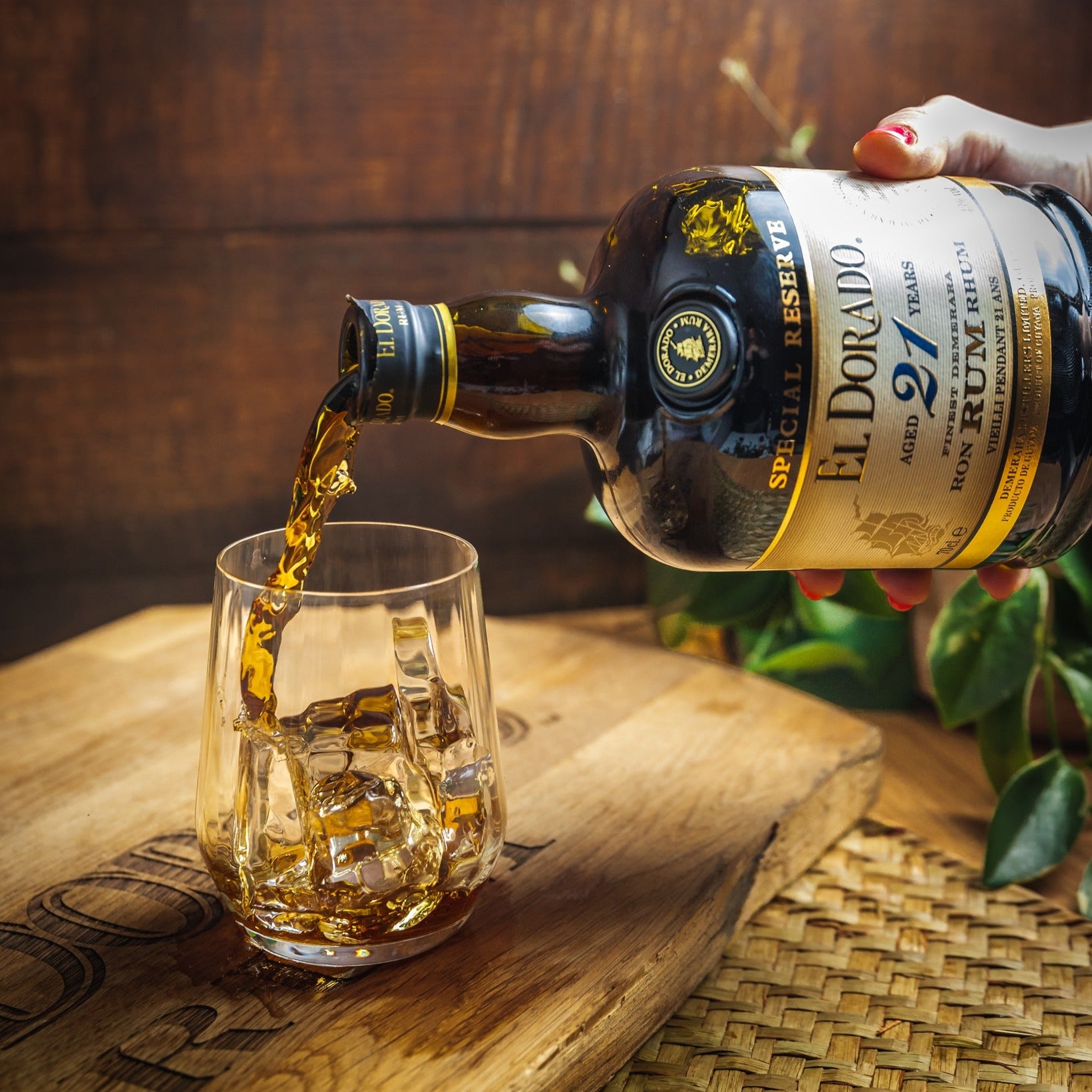 Load image into Gallery viewer, El Dorado 21YO rum being poured into a glass with a wooden barrel and plant in the background