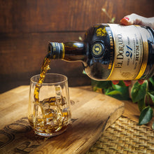 Load image into Gallery viewer, El Dorado 21YO rum being poured into a glass with a wooden barrel and plant in the background