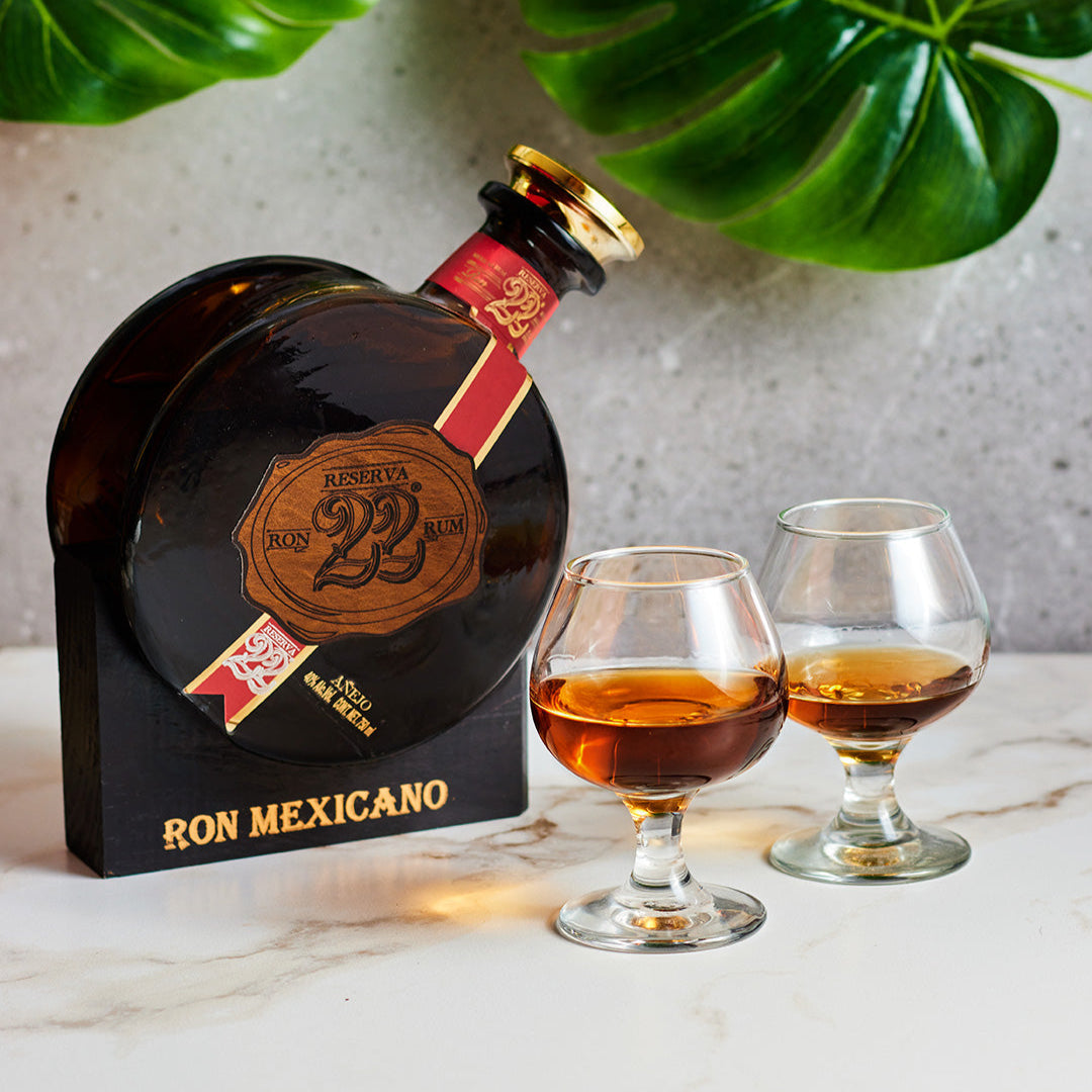 Load image into Gallery viewer, Ron Mexicano rum bottle with two glasses of rum on a marble surface with green leaves in the background.