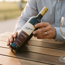 Load image into Gallery viewer, Person holding a bottle of Elderton-Command-Shiraz wine on a wooden surface with a blurred background
