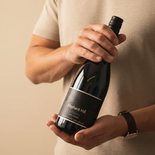 Load image into Gallery viewer, Person holding a bottle of Elephant Hill STONE Syrah wine against a beige background