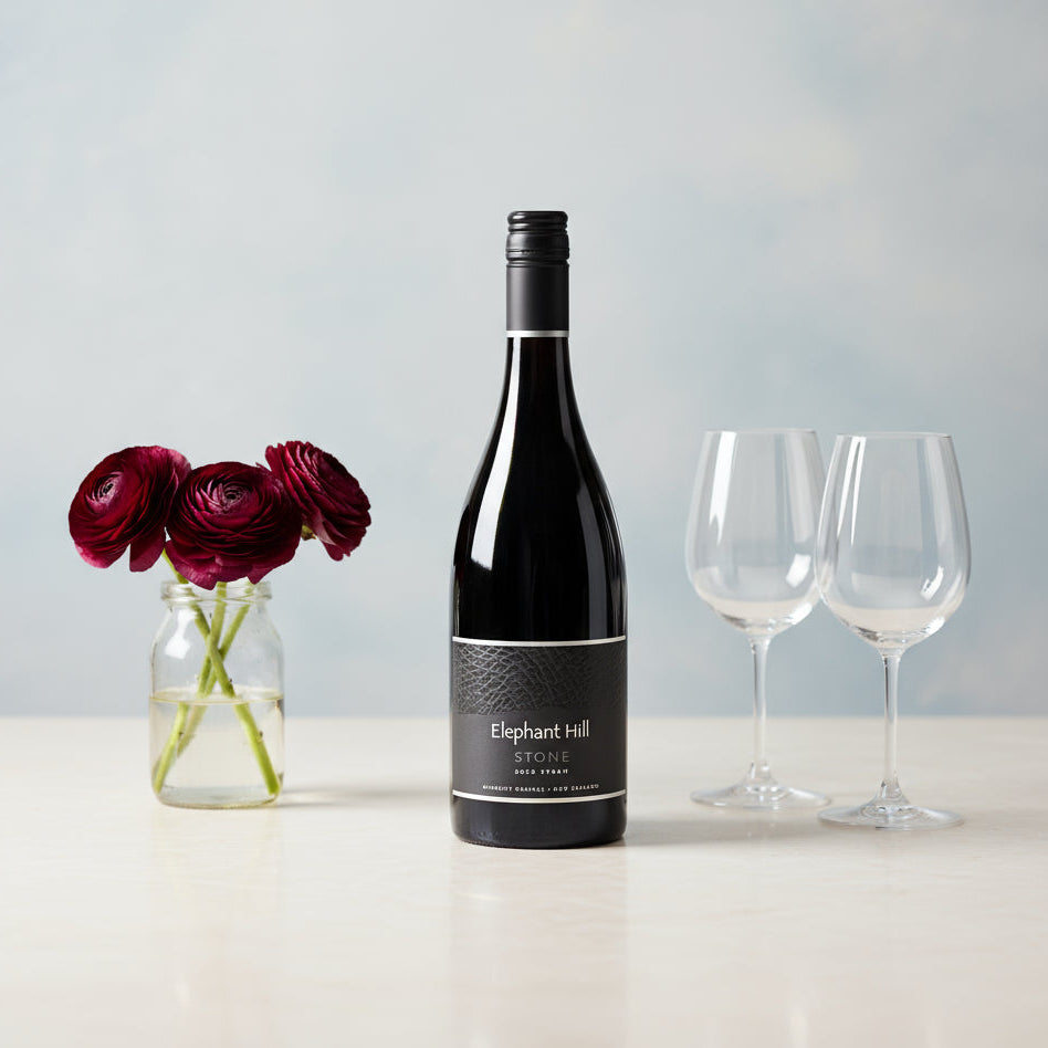 Elephant Hill STONE Syrah wine bottle with a label, two wine glasses, and a vase of red flowers on a light surface.