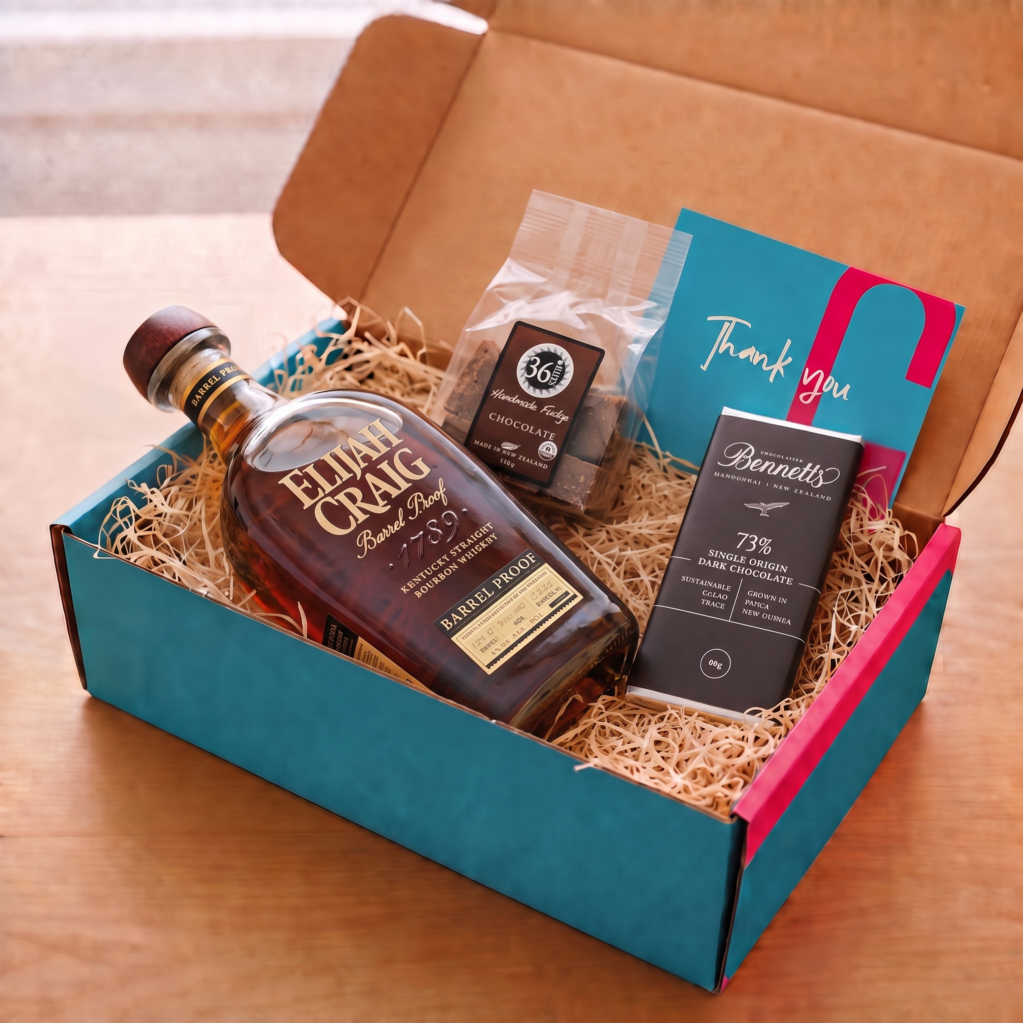 Load image into Gallery viewer, A curated gift box set on a wooden table, featuring a bottle of Elijah Craig Barrel Proof bourbon whiskey, a bar of Bennetts dark chocolate bar, a packet of 36 Degrees Chocolate fudge, a blue and pink 'Thank You' card, and shredded paper filler inside an open teal and pink COVE 27 gift box.