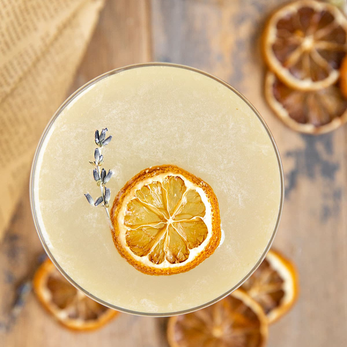 Load image into Gallery viewer, Cocktail with dried lemon slice and lavender garnish on a wooden surface.