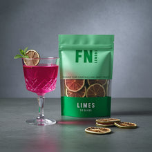 Load image into Gallery viewer, Green package of dried limes next to a pink cocktail with lime slices on a gray background