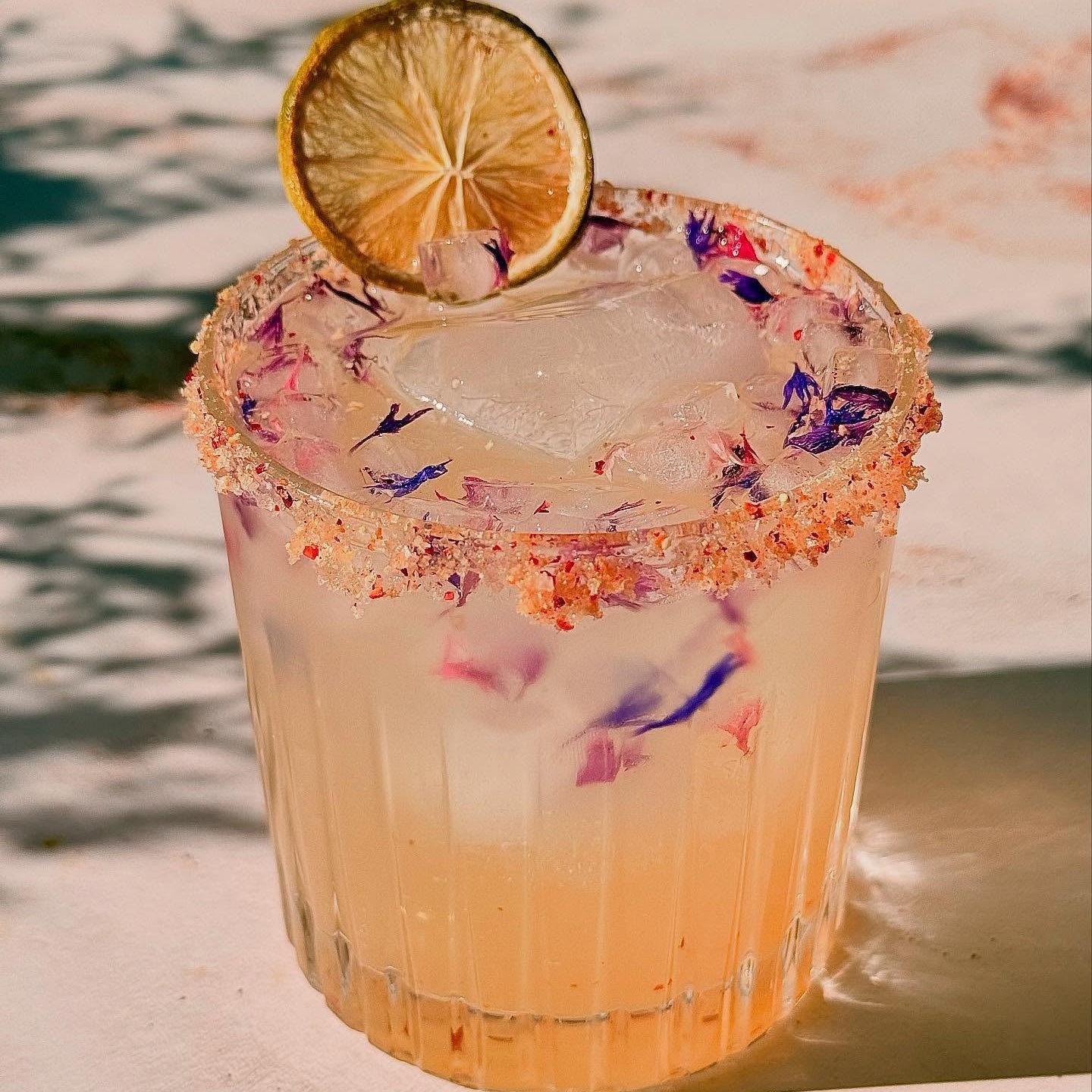 Load image into Gallery viewer, A frosted glass tumbler filled with a pale pink cocktail, topped with ice, a FN Lemon Simply Blush Edible Florets flower-rimmed edge, and a dried lime wheel garnish, set on a sunlit surface with soft shadows.