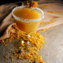 Load image into Gallery viewer, Orange cocktail with a sugared rim on a wooden surface with calendula flowers.