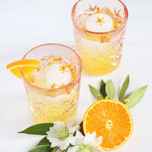 Load image into Gallery viewer, Two cocktail glasses with orange flower petals, sliced orange fruit with ice cubes and flowers, on a white background.