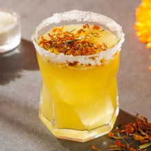 Load image into Gallery viewer, Yellow cocktail in a glass with a salt rim and floral garnish on a textured surface.