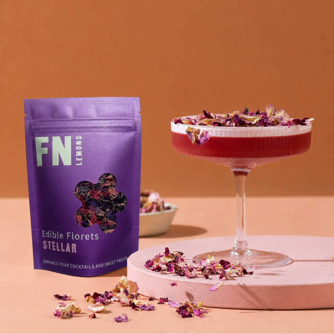Load image into Gallery viewer, A purple stand-up pouch labeled 'FN Lemons Edible Florets Stellar' stands beside a coupe glass filled with a pink-red drink topped with dried edible flower petals, on a peach studio set with a pink platform.